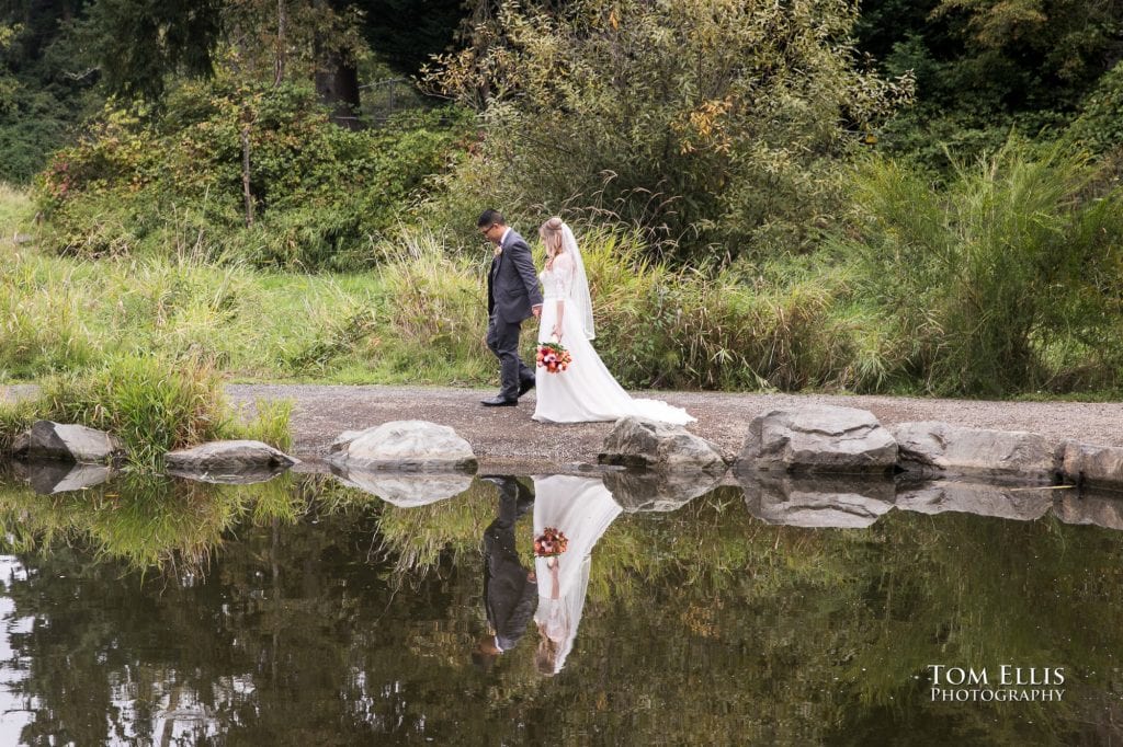 Bride and groom are reflected in the mirror-like water as they walk alongside a beautiful pond