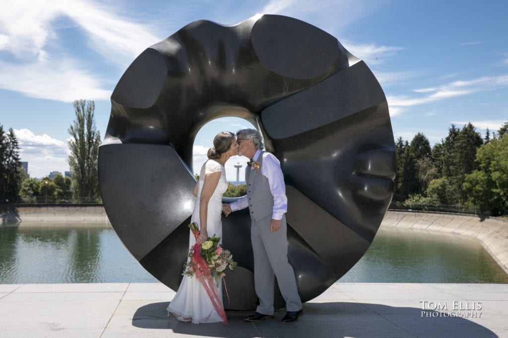 Newlywed couple kiss in front of the sculpture at Volunteer Park in Seattle, with a backdrop of downtown Seattle and the Space Needle framed by the sculpture