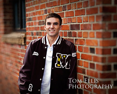 Senior boy in letterman's jacket leaning against old brick building at Luther Burbank Park