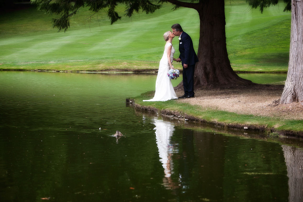 Seattle wedding photographer Tom Ellis Photography. Bride and groom share a kiss while standing along a beautiful pond, with reflection in the water