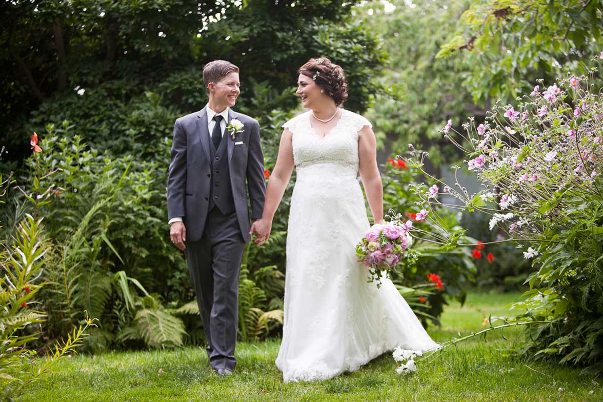 Seattle wedding photographer Tom Ellis Photography. Same-sex brides walk together holding hands through a beautiful garden