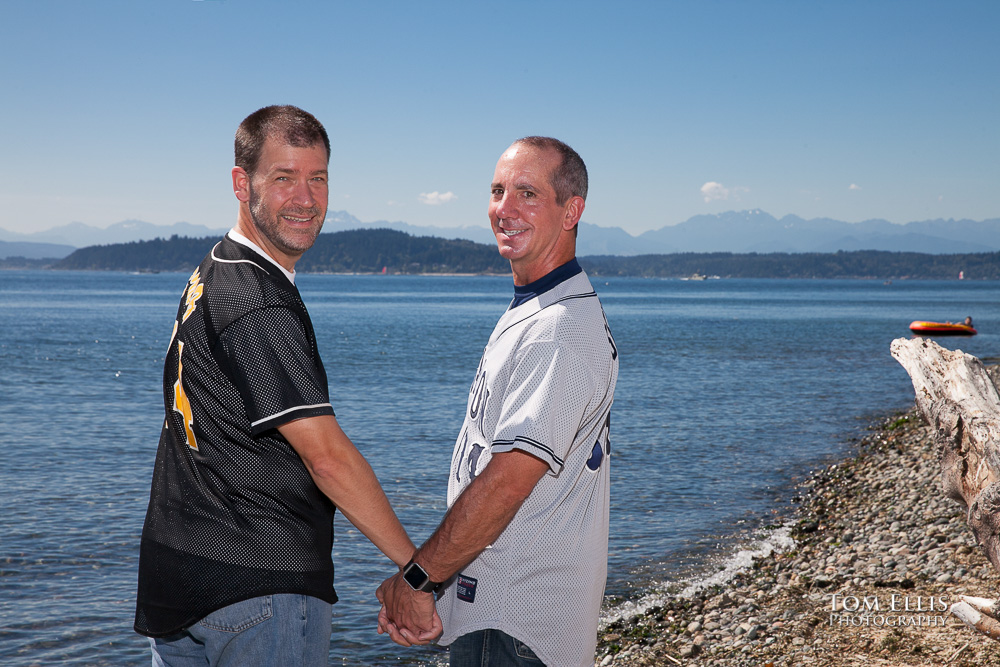 Gay couple walk hand in hand along beach, look back over shoulder at the camera