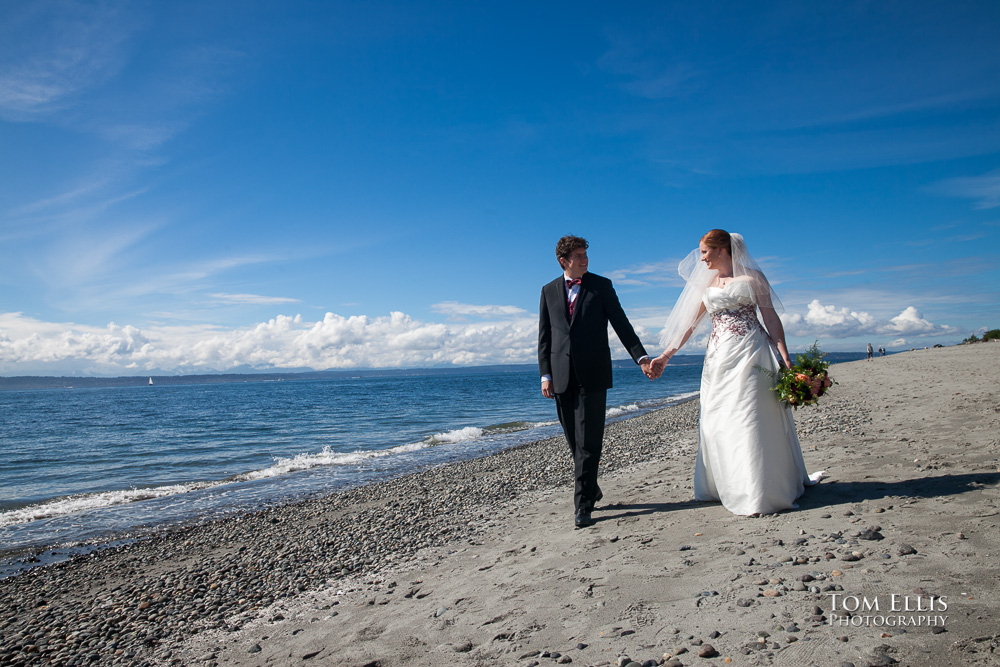 Bride and groom walk hand-in-hand along the beach under beautiful blue skies, at Golden Gardens Park in Seattle