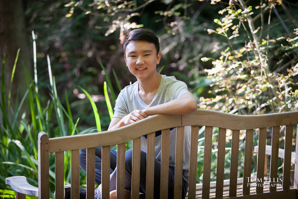 High school senior boy sits on a bench in the woods, with dramatic backlighting