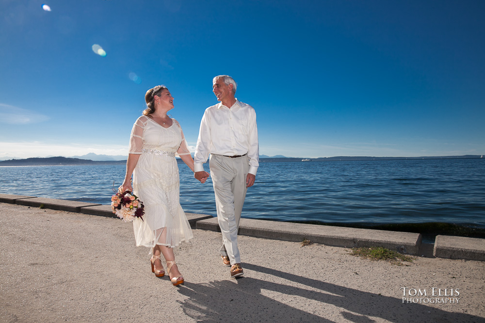 Bride and groom walk along the beachfront on Alki Point, with Puget Sound and the Olympic Mountains in the background