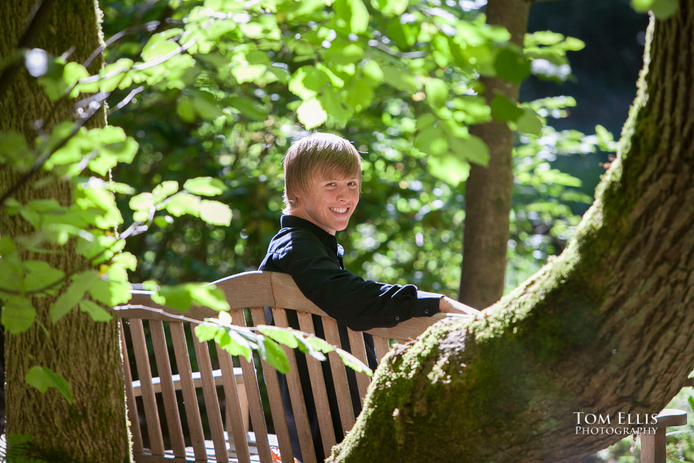 Senior boy sites on a bench surrounded by tress with beautiful backlighting highlighting him