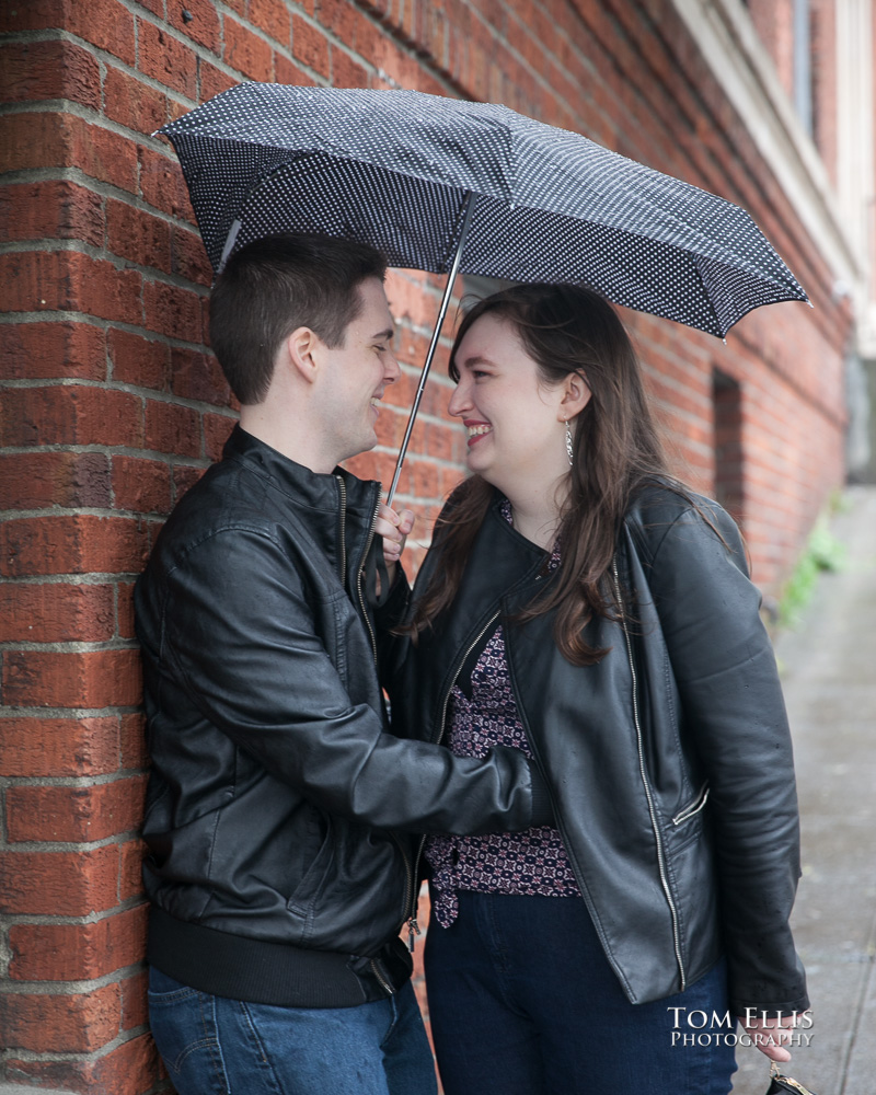 Engagement photo of couple standing under an umbrella against a brick wall in Seattle's Pike Place Market