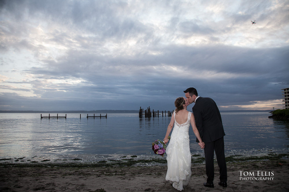 Bride and groom kiss while standing on a beautiful beach along Puget Sound, next to the Ballard Bay Club in Seattle