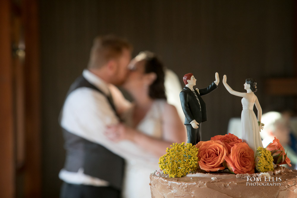 Close up photo of the wedding cake with funny topper, with bride and groom kissing in the background