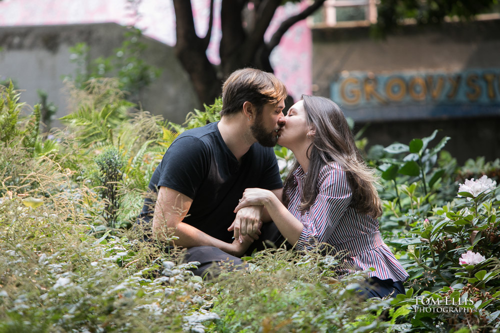 Engaged couple share a kiss while sitting in the shrubbery during their Seattle engagement photo session