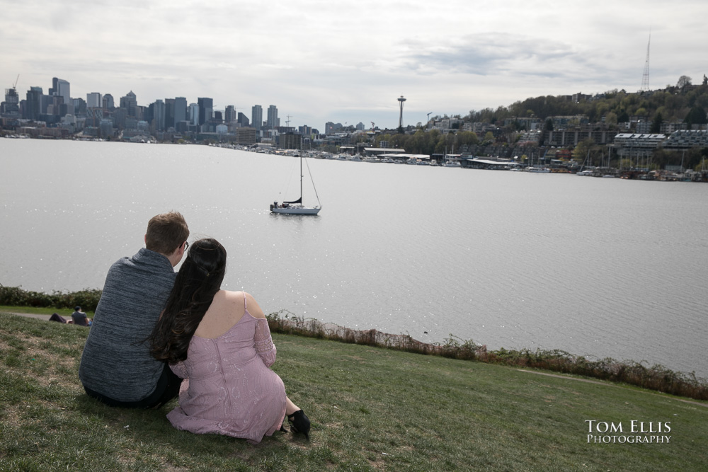 Yoshimi and Jake sit together and look across Lake Union to downtown Seattle during their engagement photo session at Gas Works Park