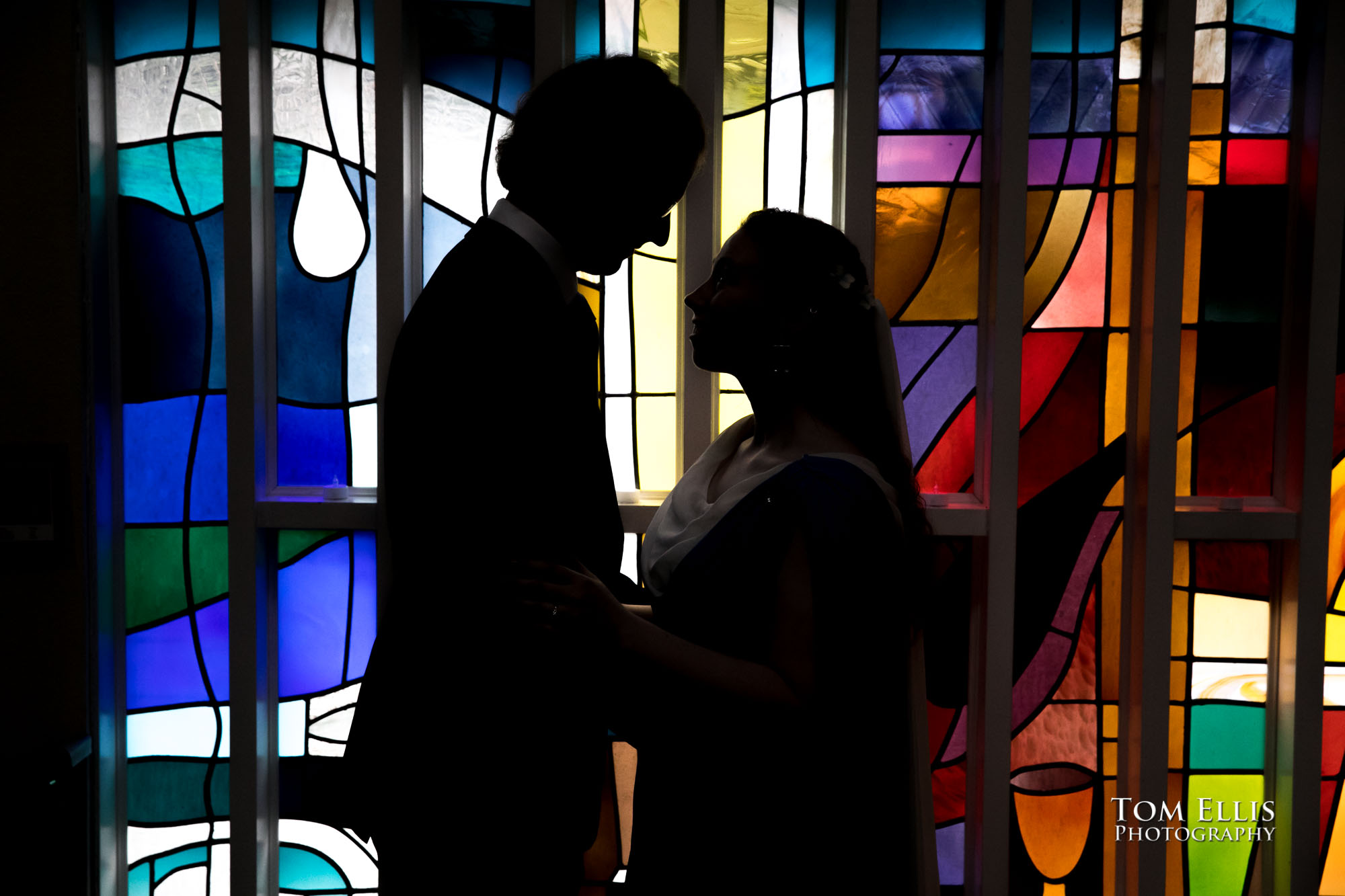 Bride Dorothy Ann and groom Mathew in silhouette against the stain glass window before their wedding ceremony at Maple Leaf Lutheran Church. Tom Ellis Photography, Seattle wedding photographer